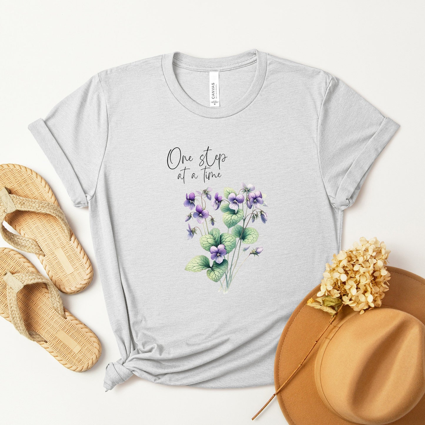 A t-shirt featuring a beautiful violet for February with the quote “One step at a time.” Perfect for February birthdays and floral art lovers. Comfortable and stylish for casual outings or celebrations. Bella+Canvas 3001 t-shirt in ash. ReadyShirtAim.com