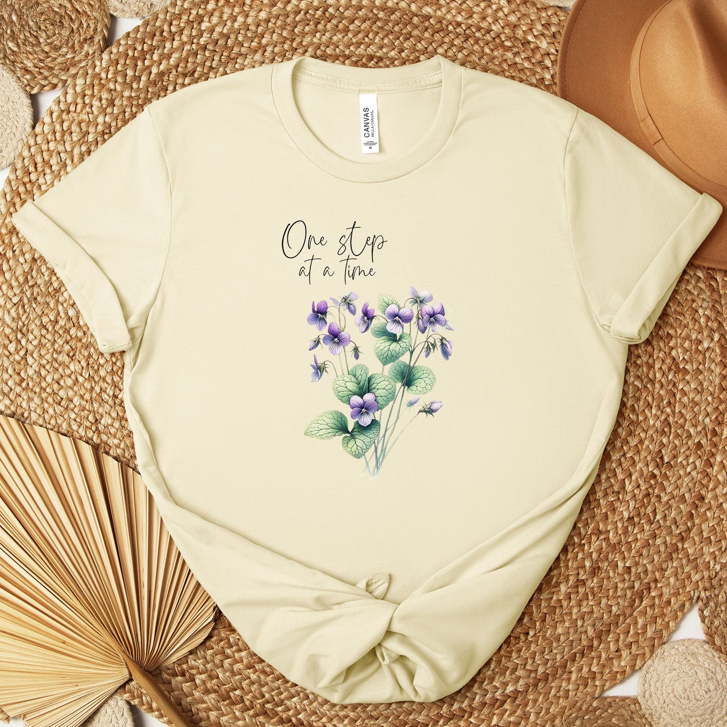 A t-shirt featuring a beautiful violet for February with the quote “One step at a time.” Perfect for February birthdays and floral art lovers. Comfortable and stylish for casual outings or celebrations. Bella+Canvas 3001 t-shirt in soft cream. ReadyShirtAim.com