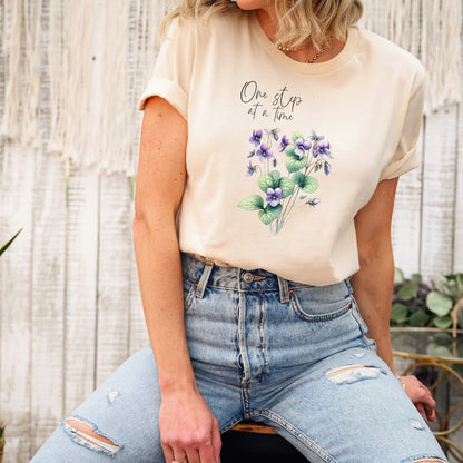 A t-shirt featuring a beautiful violet for February with the quote “One step at a time.” Perfect for February birthdays and floral art lovers. Comfortable and stylish for casual outings or celebrations. Bella+Canvas 3001 t-shirt in soft cream. ReadyShirtAim.com