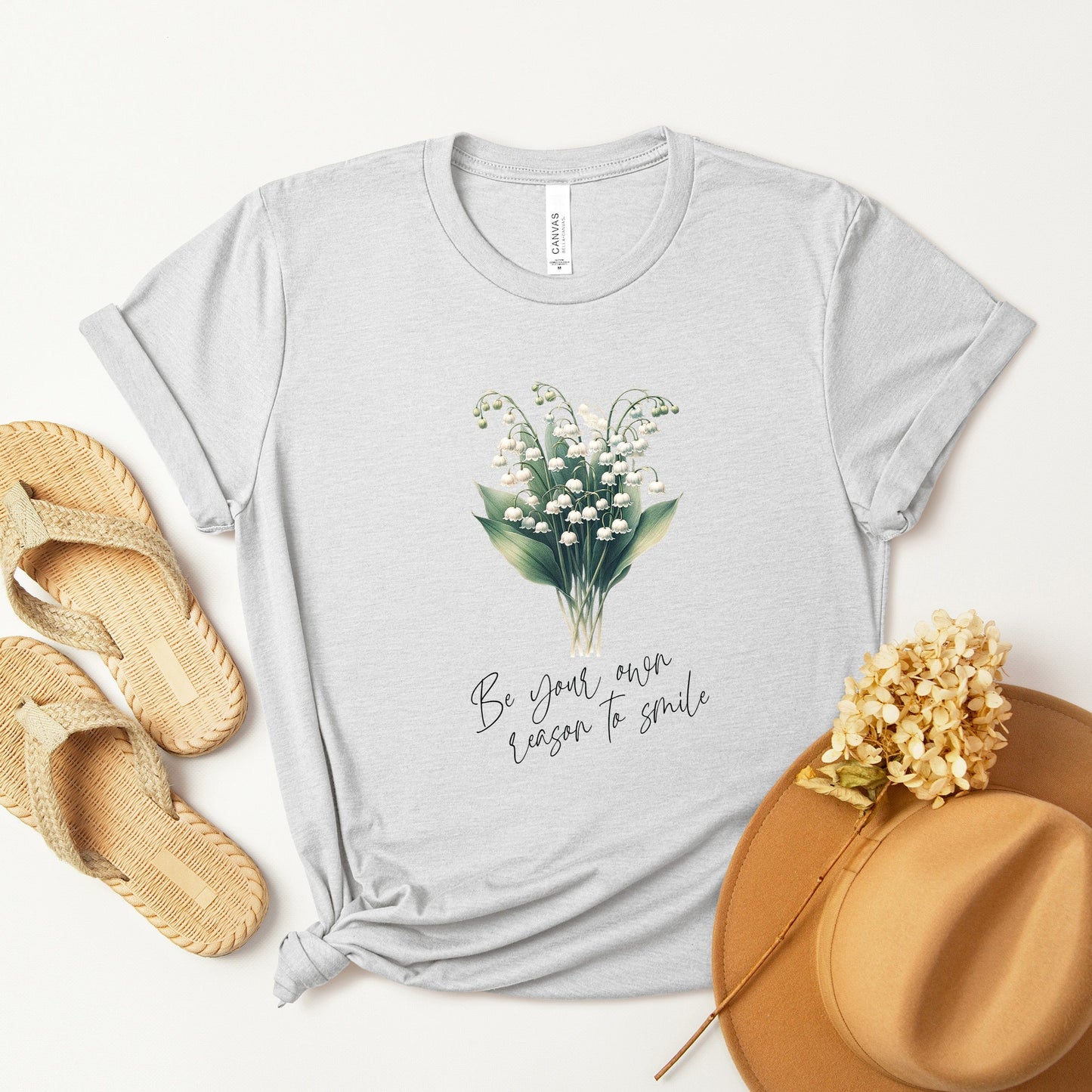 A t-shirt featuring a beautiful Lily of the Valley for May with the quote “Be your own reason to smile.” Perfect for May birthdays and floral art lovers. Comfortable and stylish for casual outings or celebrations. Bella+Canvas 3001 t-shirt in ash. ReadyShirtAim.com