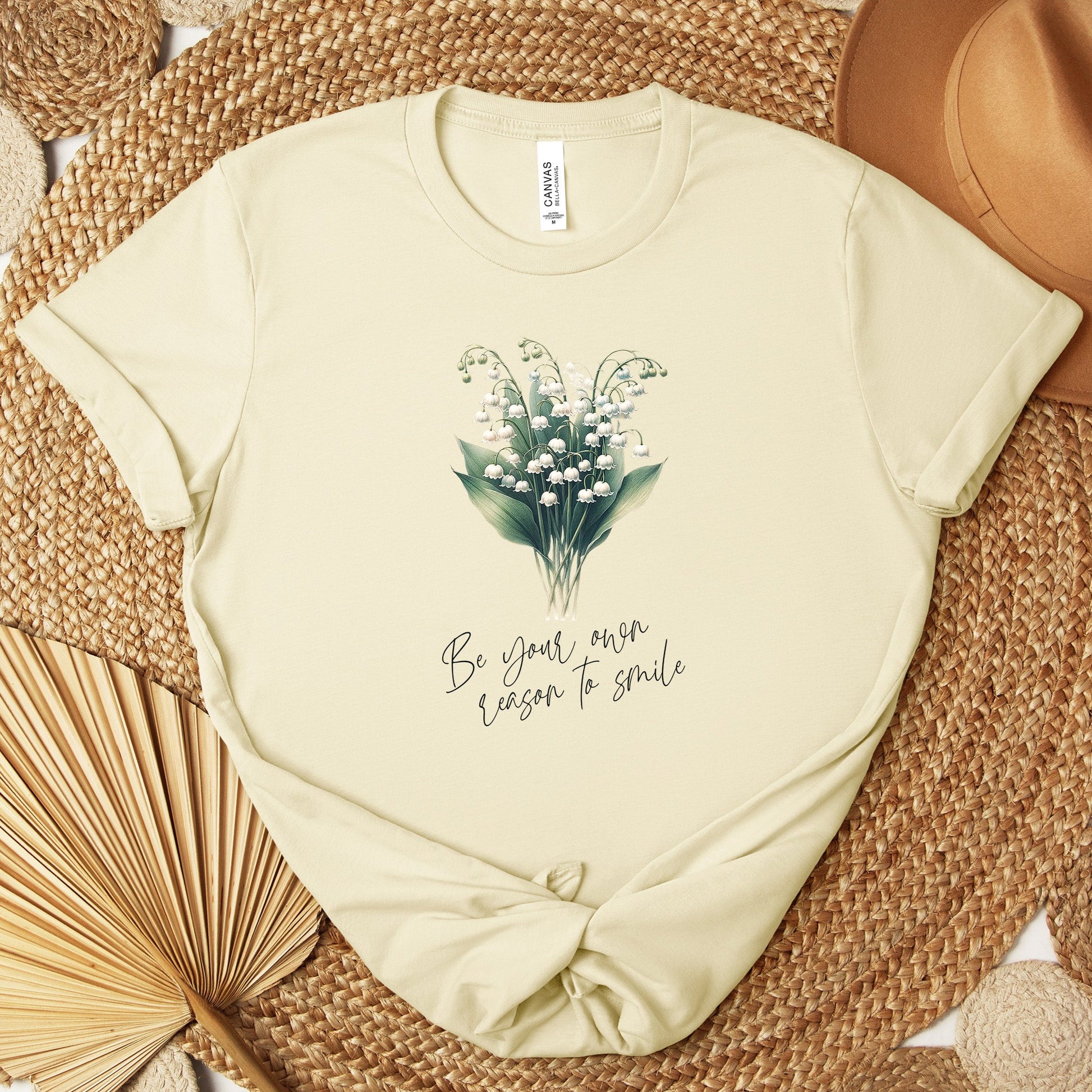 A t-shirt featuring a beautiful Lily of the Valley for May with the quote “Be your own reason to smile.” Perfect for May birthdays and floral art lovers. Comfortable and stylish for casual outings or celebrations. Bella+Canvas 3001 t-shirt in natural. ReadyShirtAim.com