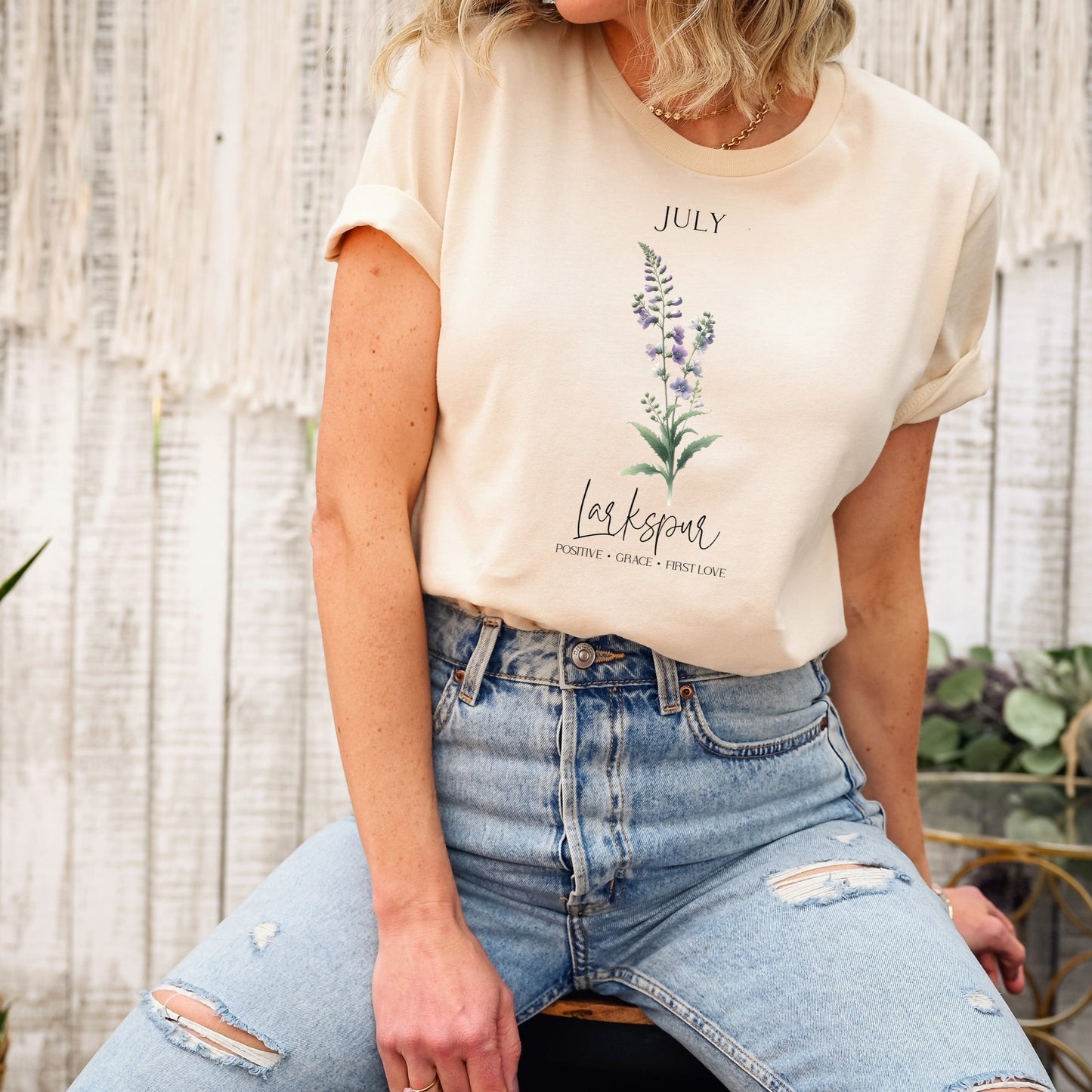 A t-shirt featuring a beautiful larkspur for July with traits Positive, Grace, First Love. Perfect for July birthdays and floral art lovers. Comfortable and stylish for casual outings or celebrations. Bella+Canvas 3001 t-shirt in soft cream. ReadyShirtAim.com