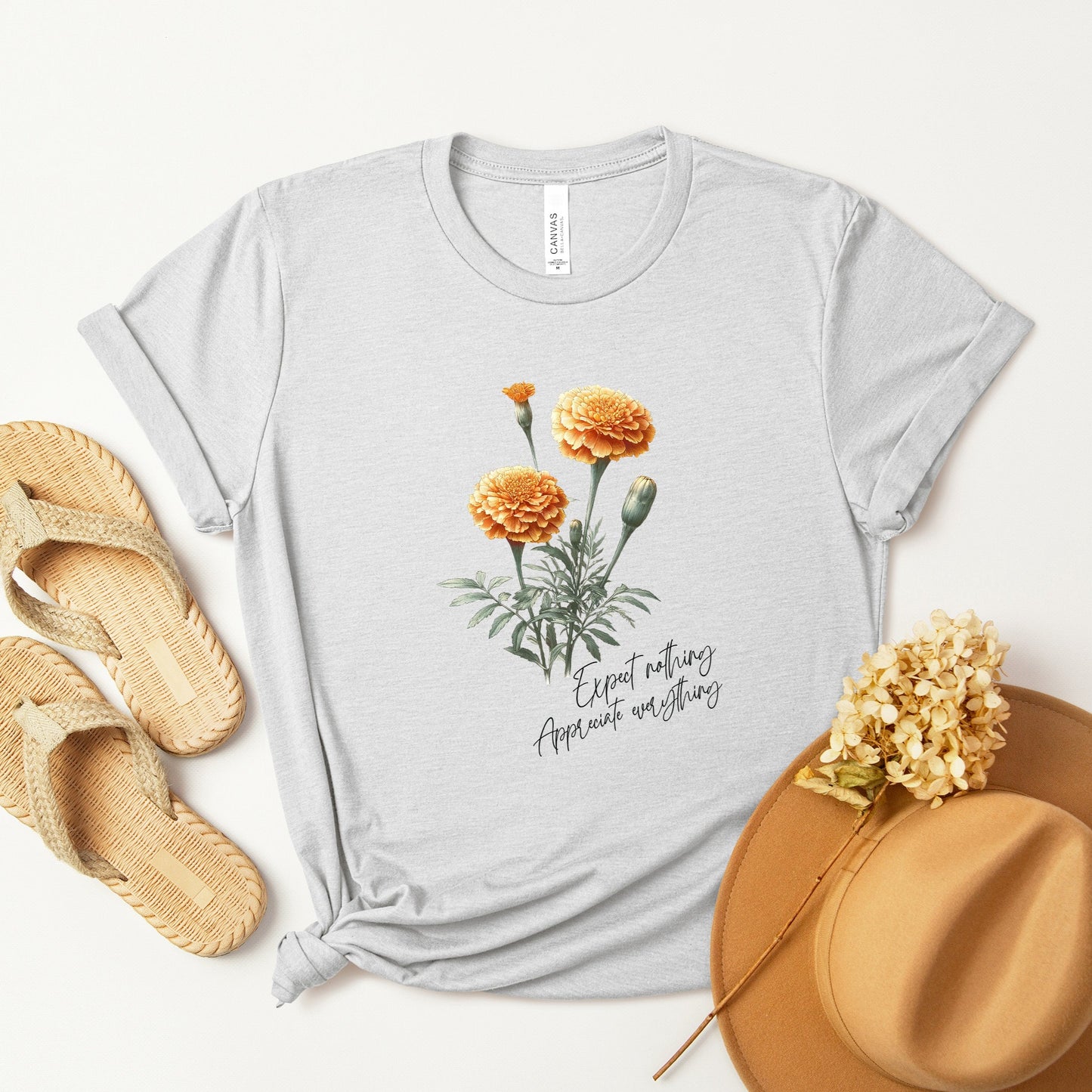 A t-shirt featuring a beautiful marigold for October with the quote “Expect nothing, appreciate everything.” Perfect for October birthdays and floral art lovers. Comfortable and stylish for casual outings or celebrations. Bella+Canvas 3001 t-shirt in ash. ReadyShirtAim.com