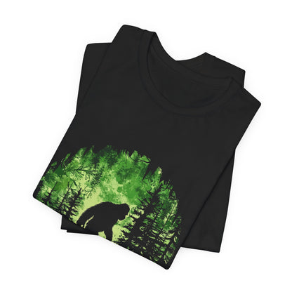 A t-shirt featuring Bigfoot in his natural habitat in shades of green, perfect for cryptid enthusiasts, outdoor adventurers, and mystery lovers. Comfortable and stylish for casual outings or cryptid conventions. Bella+Canvas 3001 t-shirt in black. ReadyShirtAim.com