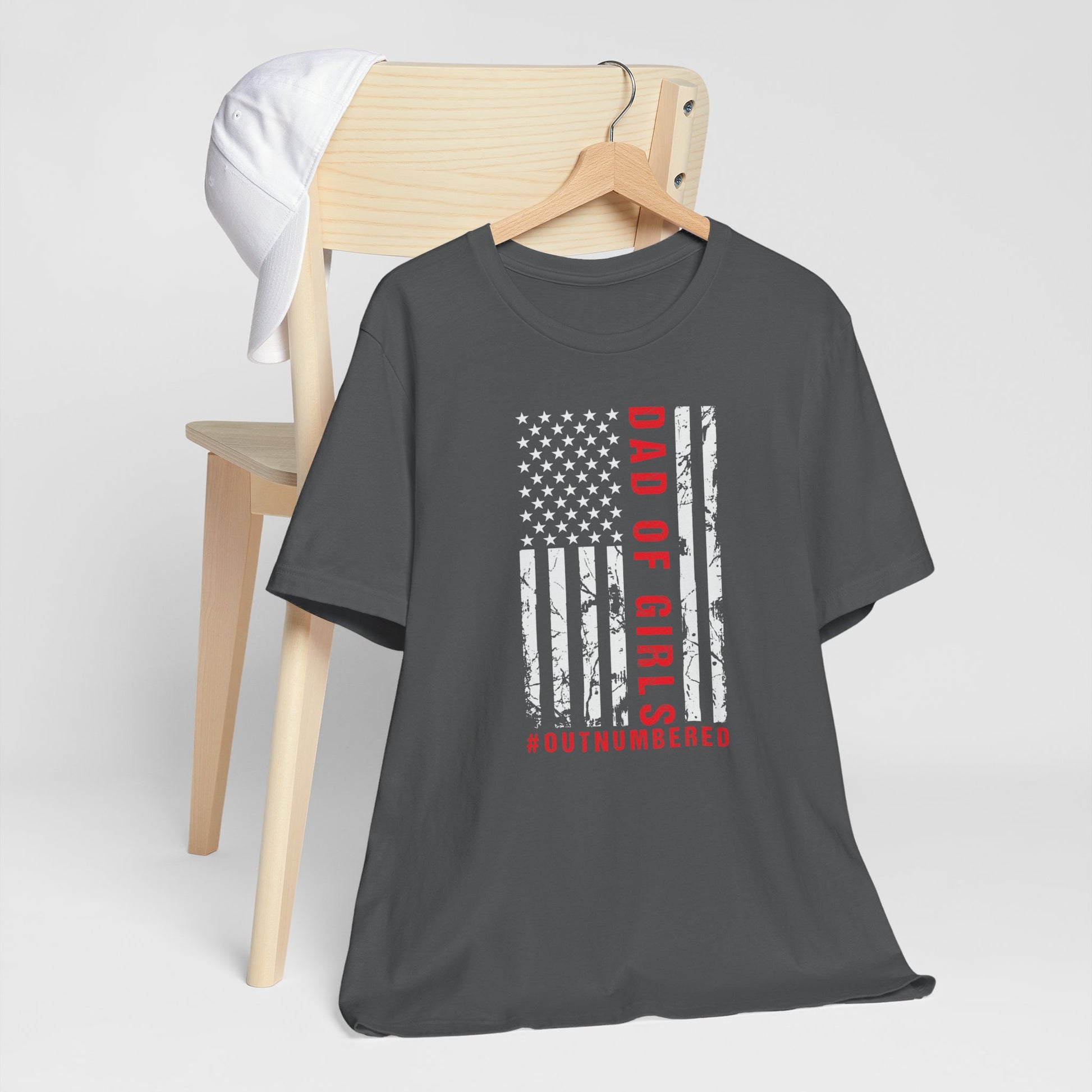 A dark-colored t-shirt featuring a grungy American flag design with the text "Dad of girls #outnumbered" in white and red. Perfect for Father's Day, Fourth of July, and patriotic dads who love humor. Comfortable and stylish for casual wear. Bella+Canvas 3001 t-shirt in asphalt. ReadyShirtAim.com