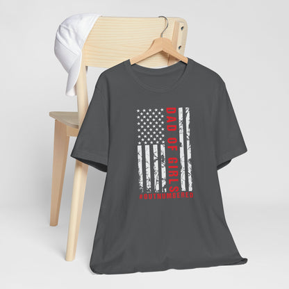 A dark-colored t-shirt featuring a grungy American flag design with the text "Dad of girls #outnumbered" in white and red. Perfect for Father's Day, Fourth of July, and patriotic dads who love humor. Comfortable and stylish for casual wear. Bella+Canvas 3001 t-shirt in asphalt. ReadyShirtAim.com