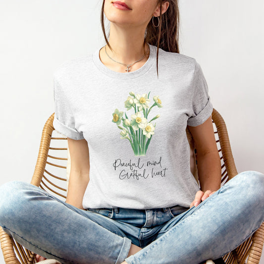 A t-shirt featuring a beautiful narcissus for December with the quote “Peaceful mind, grateful heart.” Perfect for December birthdays and floral art lovers. Comfortable and stylish for casual outings or celebrations. Bella+Canvas 3001 t-shirt in ash. ReadyShirtAim.com