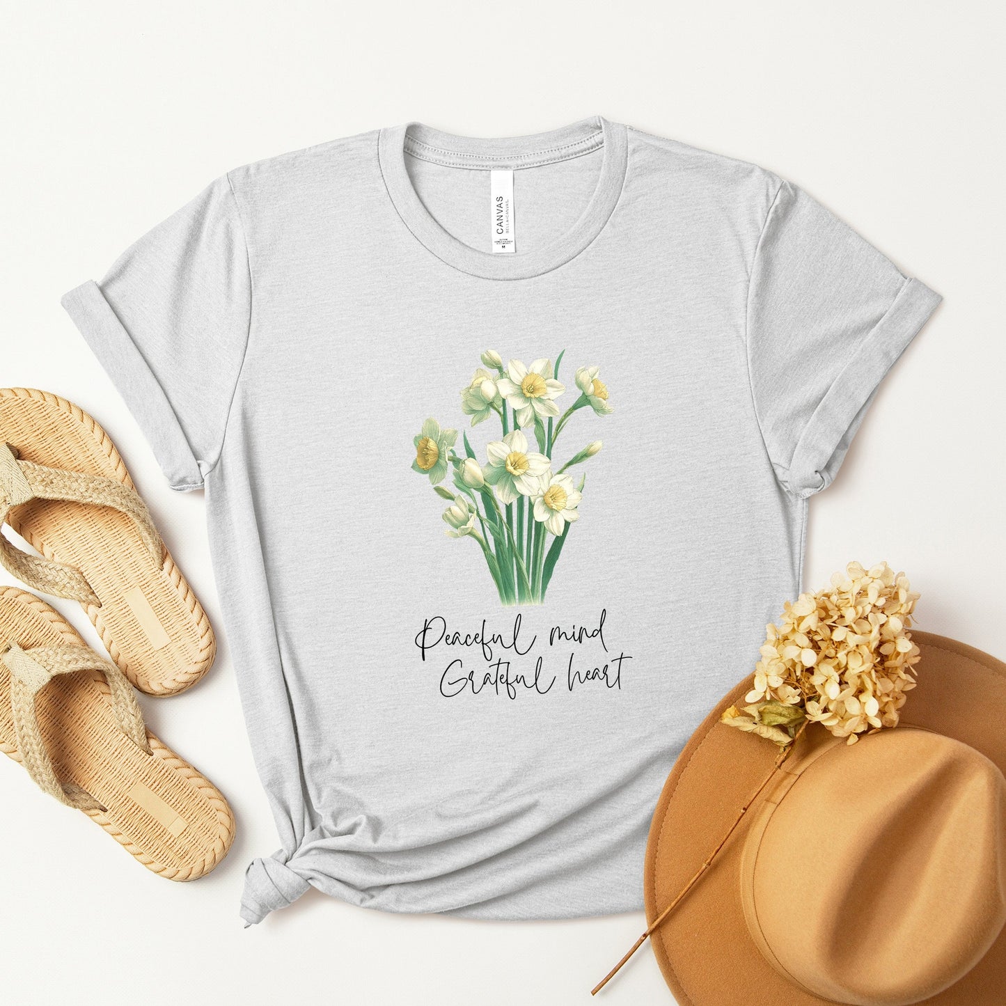 A t-shirt featuring a beautiful narcissus for December with the quote “Peaceful mind, grateful heart.” Perfect for December birthdays and floral art lovers. Comfortable and stylish for casual outings or celebrations. Bella+Canvas 3001 t-shirt in ash. ReadyShirtAim.com