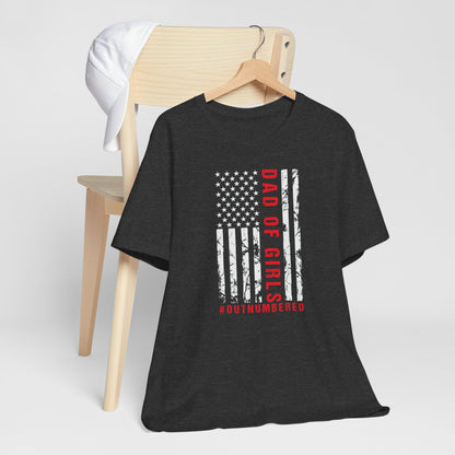 A dark-colored t-shirt featuring a grungy American flag design with the text "Dad of girls #outnumbered" in white and red. Perfect for Father's Day, Fourth of July, and patriotic dads who love humor. Comfortable and stylish for casual wear. Bella+Canvas 3001 t-shirt in dark grey heather. ReadyShirtAim.com