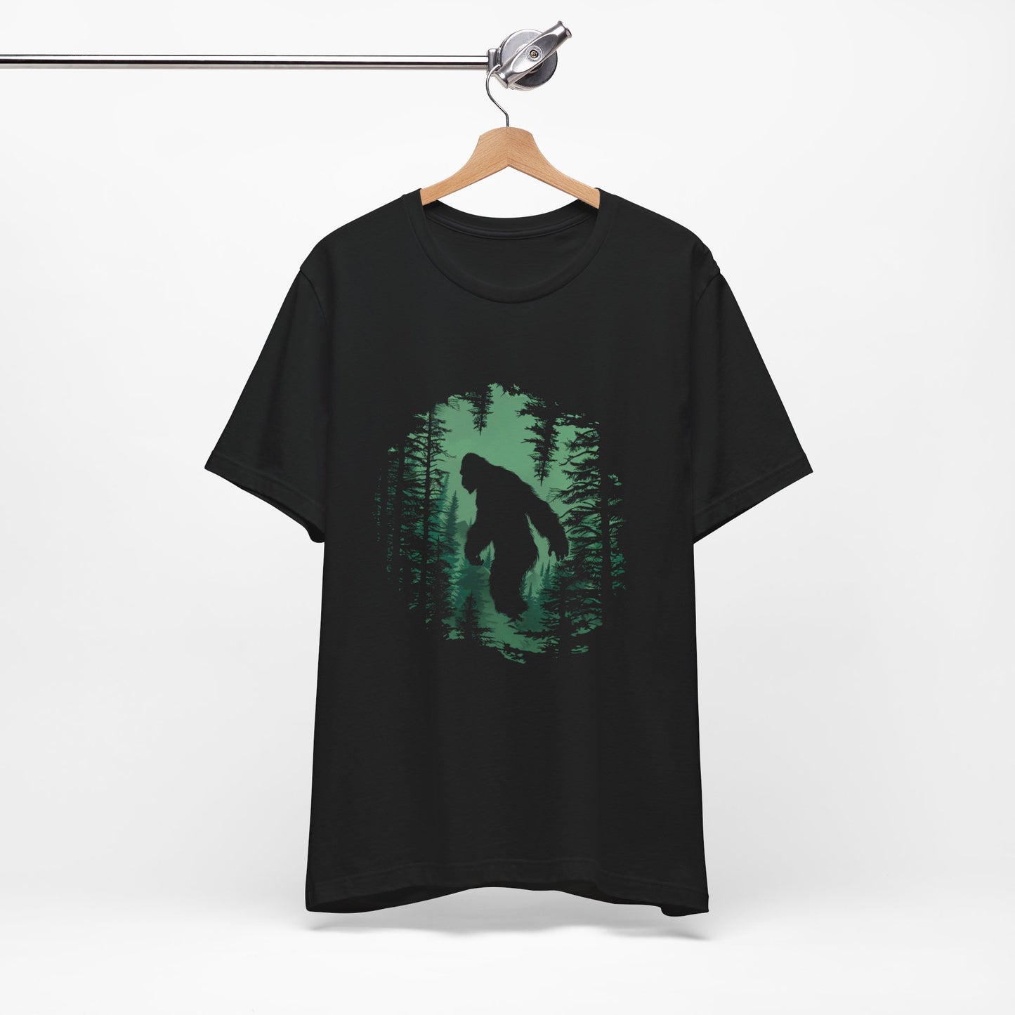 A t-shirt featuring a silhouette of Bigfoot walking in the woods in shades of green, perfect for cryptid enthusiasts, outdoor adventurers, and mystery lovers. Comfortable and stylish for casual outings or cryptid conventions. Bella+Canvas 3001 t-shirt in black. ReadyShirtAim.com