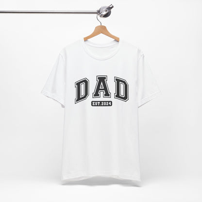 A stylish light colored t-shirt featuring the text "Dad Est. 2024" in a collegiate, sporty font, perfect for new fathers. Made from soft, breathable cotton, this shirt is an ideal gift for Father's Day or to celebrate a newborn's arrival. Available in multiple colors and sizes. Bella+Canvas 3001 t-shirt. ReadyShirtAim.com