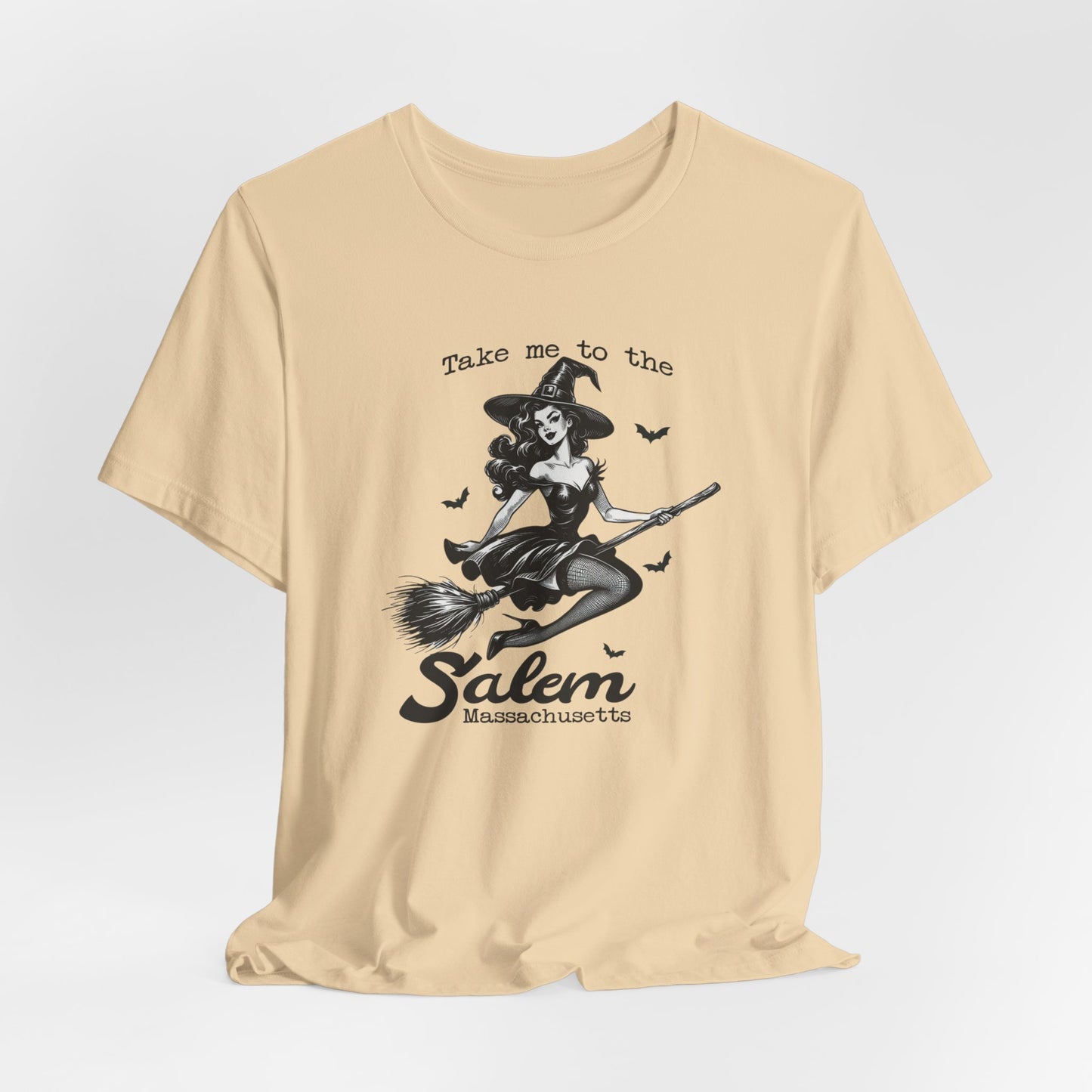 "Take Me to Salem, Massachusetts" Vintage 1950's Witch Halloween Women's T-Shirt featuring bold black text and a classic black-and-white image of a pin-up girl in a sexy witch costume flying gracefully on a broomstick with bats surrounding her, perfect for adding a retro touch with whimsical magic to your Halloween festivities. Available in multiple colors and sizes.  Bella+Canvas 3001 t-shirt in soft cream. ReadyShirtAim.com