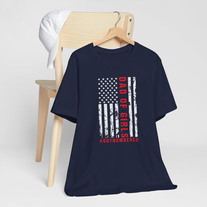 A dark-colored t-shirt featuring a grungy American flag design with the text "Dad of girls #outnumbered" in white and red. Perfect for Father's Day, Fourth of July, and patriotic dads who love humor. Comfortable and stylish for casual wear. Bella+Canvas 3001 t-shirt in navy. ReadyShirtAim.com