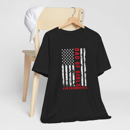 A dark-colored t-shirt featuring a grungy American flag design with the text "Dad of girls #outnumbered" in white and red. Perfect for Father's Day, Fourth of July, and patriotic dads who love humor. Comfortable and stylish for casual wear. Bella+Canvas 3001 t-shirt in black. ReadyShirtAim.com