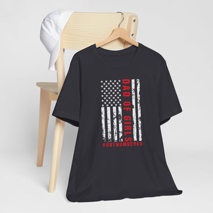 A dark-colored t-shirt featuring a grungy American flag design with the text "Dad of girls #outnumbered" in white and red. Perfect for Father's Day, Fourth of July, and patriotic dads who love humor. Comfortable and stylish for casual wear. Bella+Canvas 3001 t-shirt in dark grey. ReadyShirtAim.com