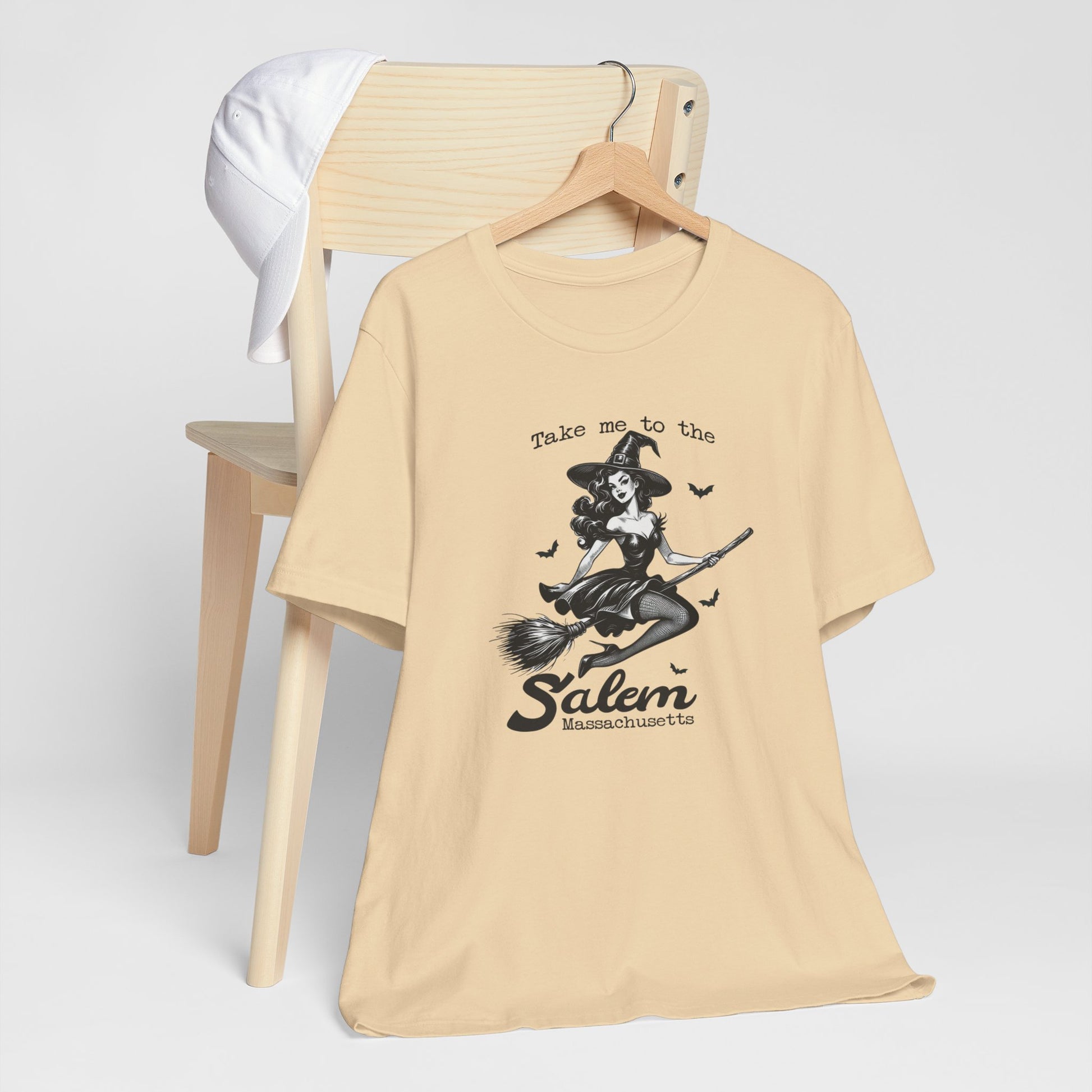 "Take Me to Salem, Massachusetts" Vintage 1950's Witch Halloween Women's T-Shirt featuring bold black text and a classic black-and-white image of a pin-up girl in a sexy witch costume flying gracefully on a broomstick with bats surrounding her, perfect for adding a retro touch with whimsical magic to your Halloween festivities. Available in multiple colors and sizes.  Bella+Canvas 3001 t-shirt in soft cream. ReadyShirtAim.com