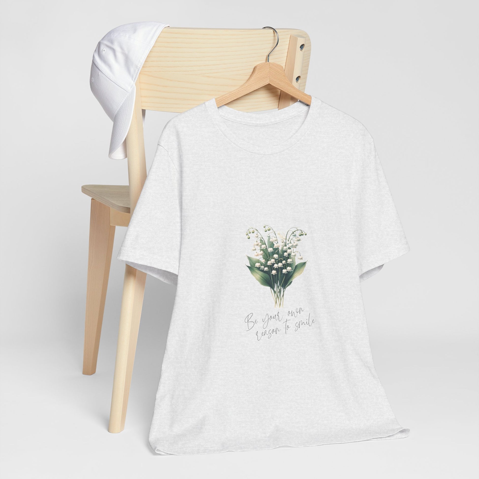 A t-shirt featuring a beautiful Lily of the Valley for May with the quote “Be your own reason to smile.” Perfect for May birthdays and floral art lovers. Comfortable and stylish for casual outings or celebrations. Bella+Canvas 3001 t-shirt in ash. ReadyShirtAim.com