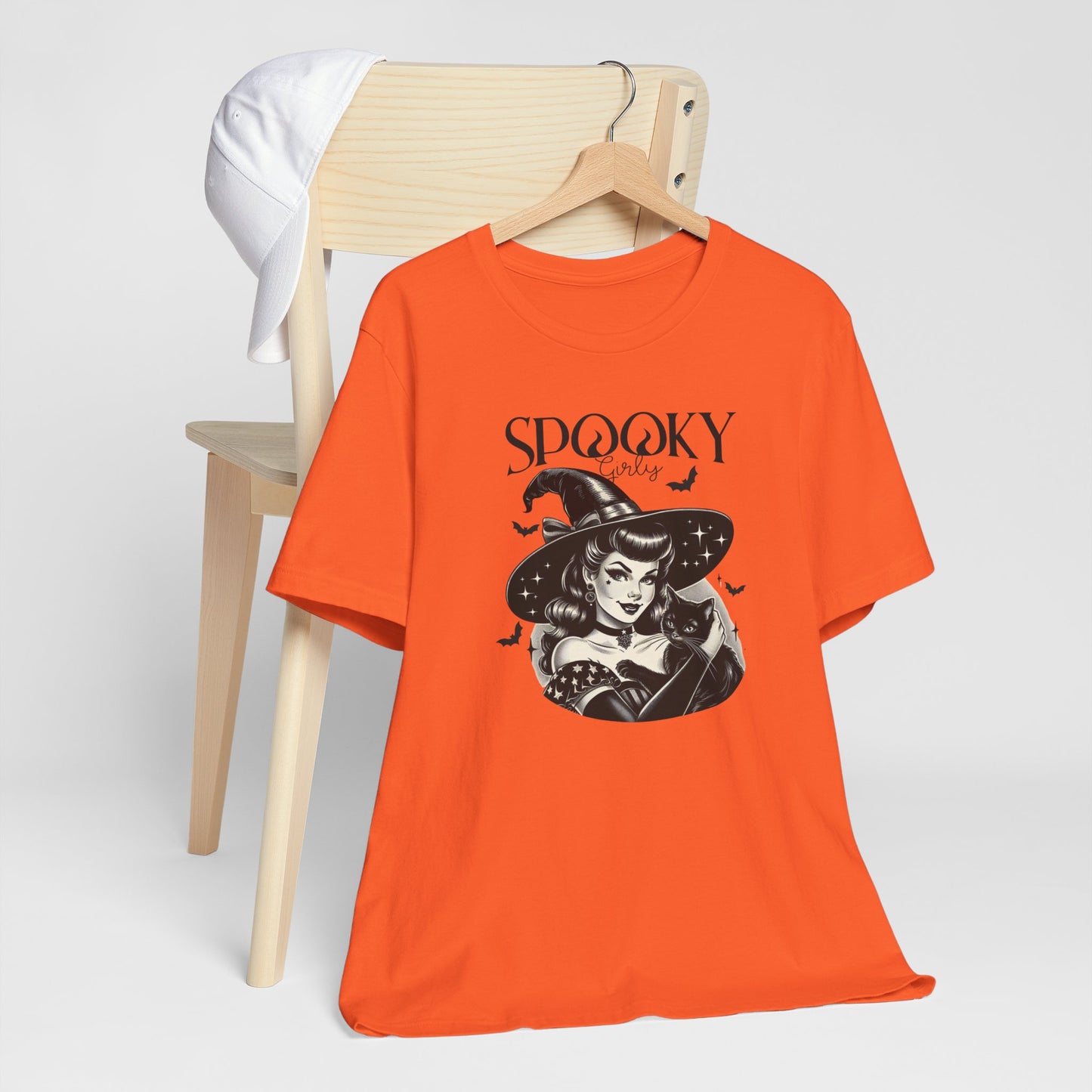 "Spooky Girly" Vintage 1950's Witch Halloween Women's T-Shirt featuring bold black text and a classic black-and-white image of a pin-up girl in a sexy witch costume, holding a black cat and smiling mischievously, perfect for adding a retro touch with playful charm to your Halloween festivities. Available in multiple colors and sizes.  Bella+Canvas 3001 t-shirt in orange. ReadyShirtAim.com