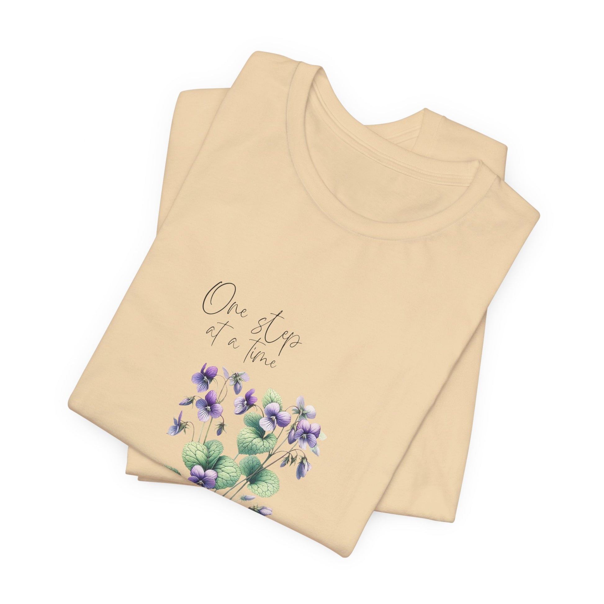 A t-shirt featuring a beautiful violet for February with the quote “One step at a time.” Perfect for February birthdays and floral art lovers. Comfortable and stylish for casual outings or celebrations. Bella+Canvas 3001 t-shirt in soft cream. ReadyShirtAim.com