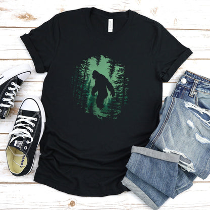 A t-shirt featuring a silhouette of Bigfoot walking in the woods in shades of green, perfect for cryptid enthusiasts, outdoor adventurers, and mystery lovers. Comfortable and stylish for casual outings or cryptid conventions. Bella+Canvas 3001 t-shirt in black. ReadyShirtAim.com