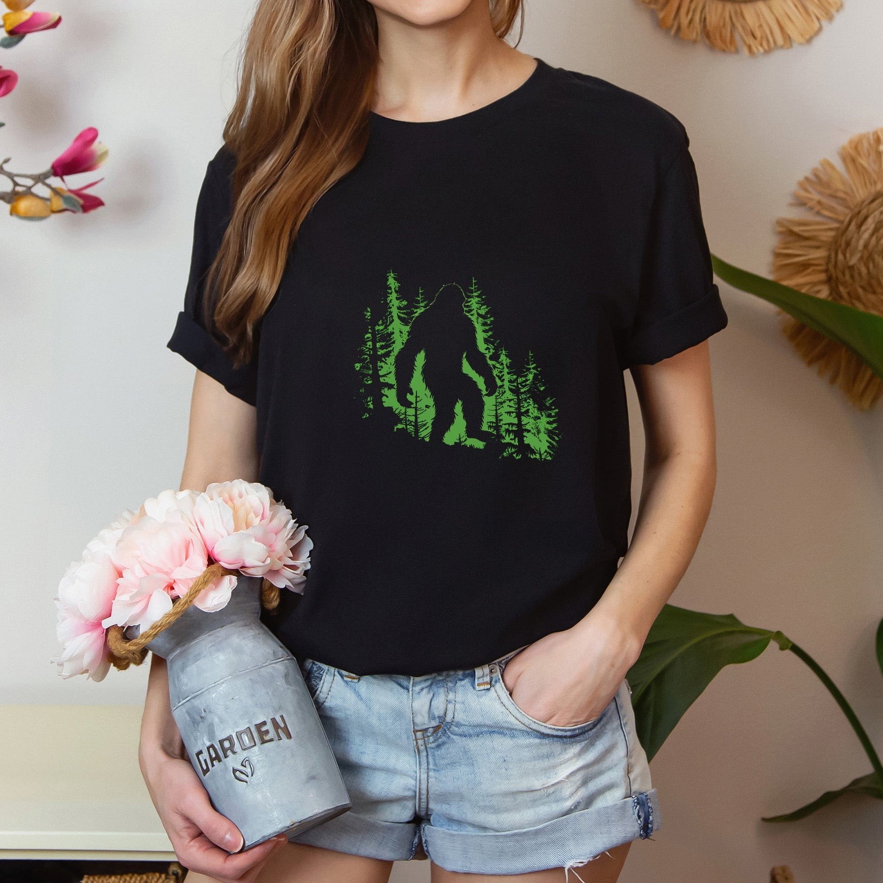 A t-shirt featuring a silhouette of Bigfoot walking in the woods in shades of green, perfect for cryptid enthusiasts, outdoor adventurers, and mystery lovers. Comfortable and stylish for casual outings or cryptid conventions. Bella+Canvas 3001 t-shirt in black. ReadyShirtAim.com