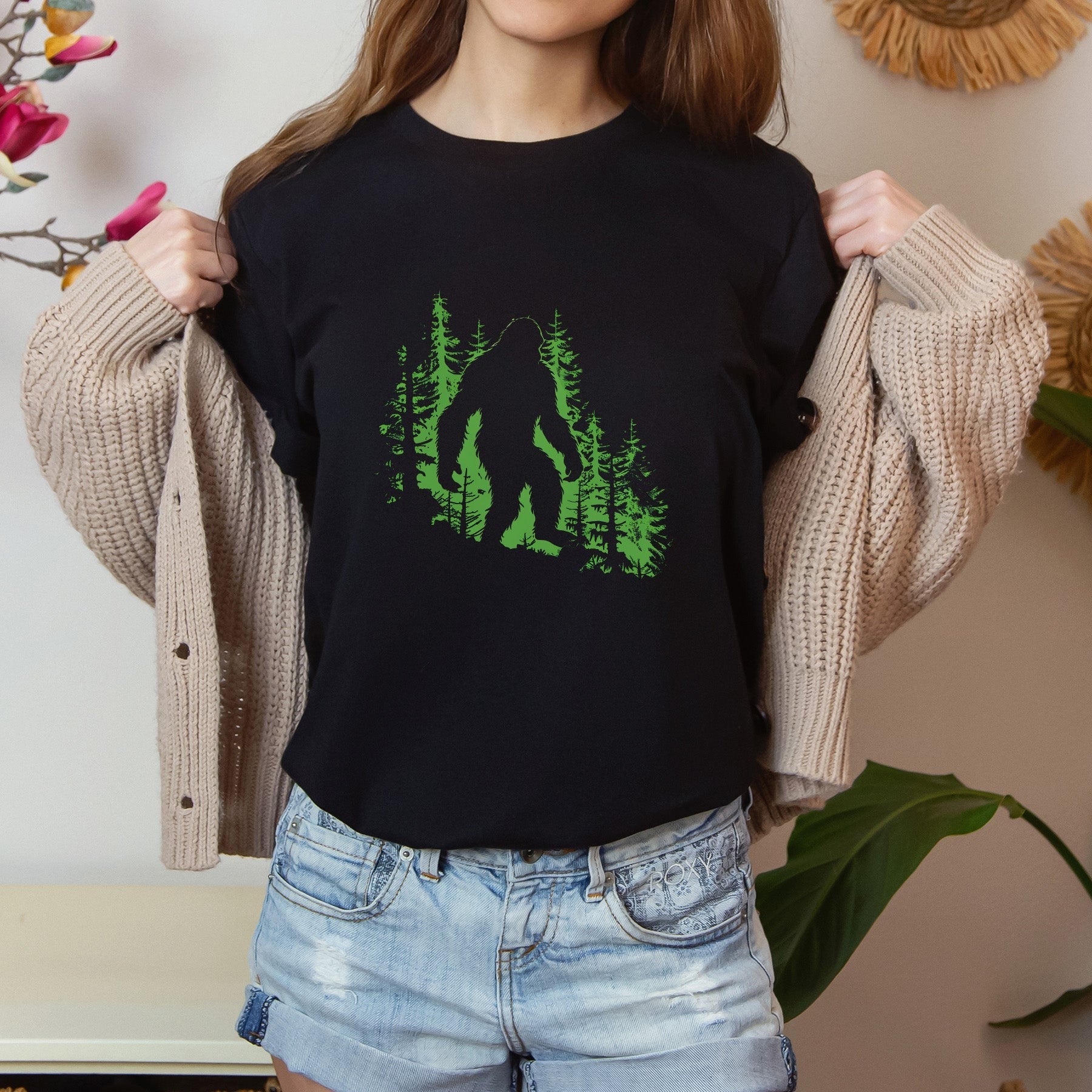 A t-shirt featuring a silhouette of Bigfoot walking in the woods in shades of green, perfect for cryptid enthusiasts, outdoor adventurers, and mystery lovers. Comfortable and stylish for casual outings or cryptid conventions. Bella+Canvas 3001 t-shirt in black. ReadyShirtAim.com