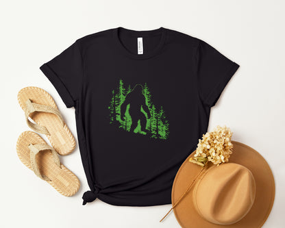 A t-shirt featuring a silhouette of Bigfoot walking in the woods in shades of green, perfect for cryptid enthusiasts, outdoor adventurers, and mystery lovers. Comfortable and stylish for casual outings or cryptid conventions. Bella+Canvas 3001 t-shirt in black. ReadyShirtAim.com