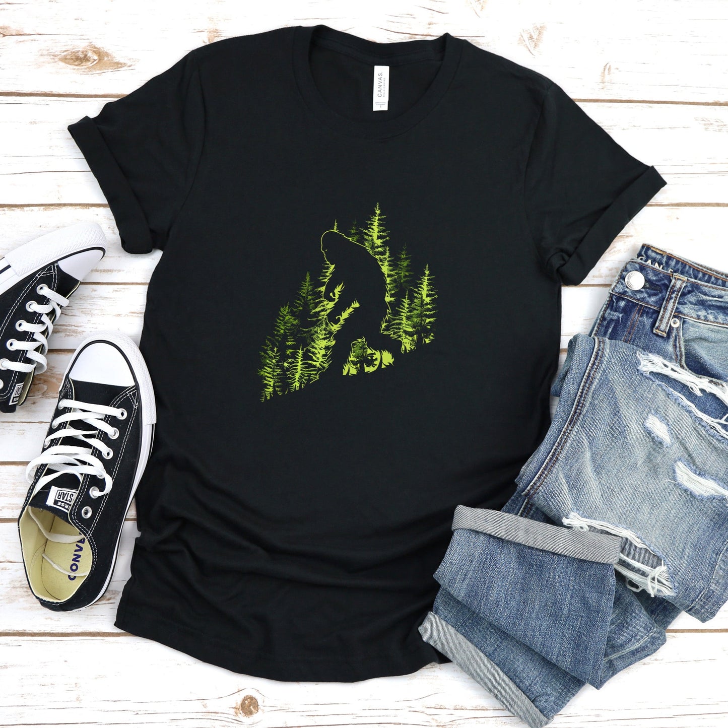 A t-shirt featuring an artistic depiction of a Bigfoot encounter, perfect for cryptid enthusiasts, outdoor adventurers, and mystery lovers. Comfortable and stylish for casual outings or cryptid conventions. Bella+Canvas 3001 t-shirt in black. ReadyShirtAim.com