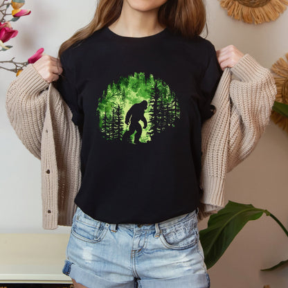 A t-shirt featuring Bigfoot in his natural habitat in shades of green, perfect for cryptid enthusiasts, outdoor adventurers, and mystery lovers. Comfortable and stylish for casual outings or cryptid conventions. Bella+Canvas 3001 t-shirt in black. ReadyShirtAim.com