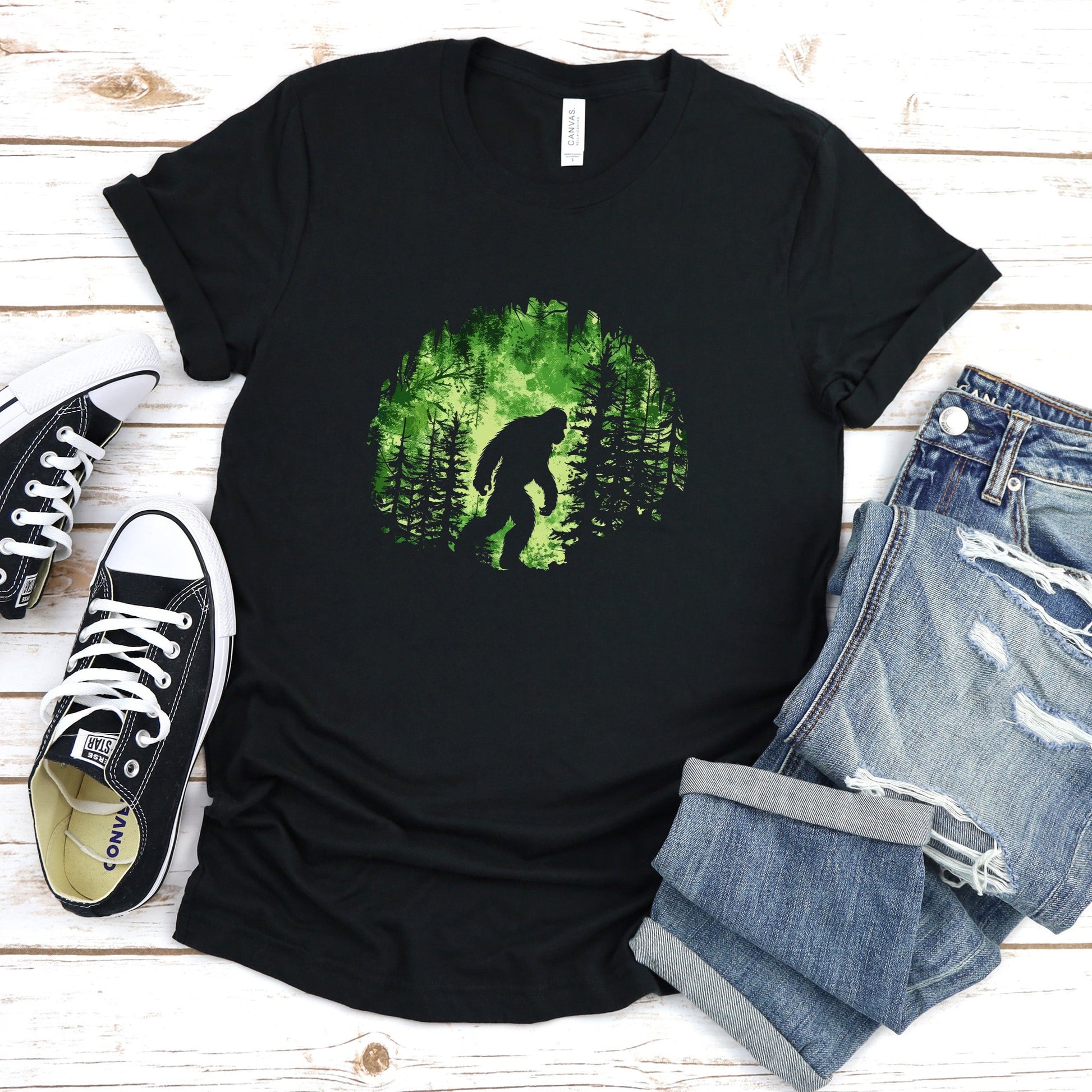 A t-shirt featuring Bigfoot in his natural habitat in shades of green, perfect for cryptid enthusiasts, outdoor adventurers, and mystery lovers. Comfortable and stylish for casual outings or cryptid conventions. Bella+Canvas 3001 t-shirt in black. ReadyShirtAim.com