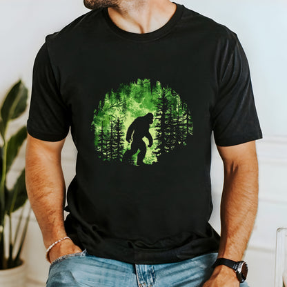 A t-shirt featuring Bigfoot in his natural habitat in shades of green, perfect for cryptid enthusiasts, outdoor adventurers, and mystery lovers. Comfortable and stylish for casual outings or cryptid conventions. Bella+Canvas 3001 t-shirt in black. ReadyShirtAim.com