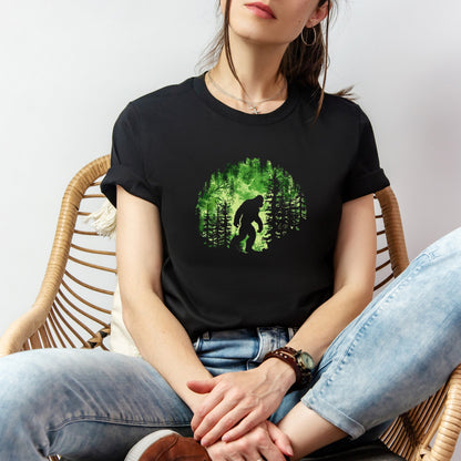 A t-shirt featuring Bigfoot in his natural habitat in shades of green, perfect for cryptid enthusiasts, outdoor adventurers, and mystery lovers. Comfortable and stylish for casual outings or cryptid conventions. Bella+Canvas 3001 t-shirt in black. ReadyShirtAim.com