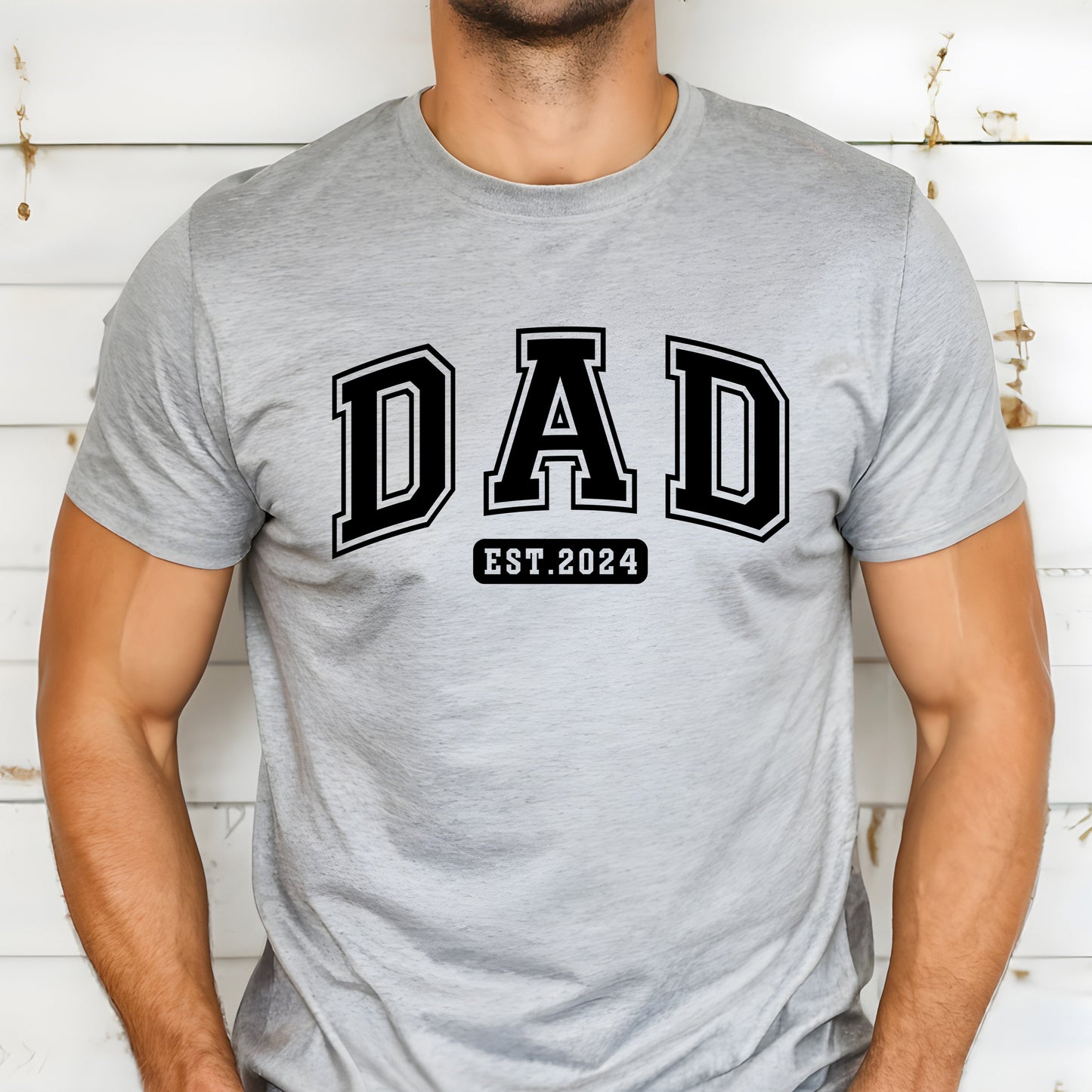 A stylish light colored t-shirt featuring the text "Dad Est. 2024" in a collegiate, sporty font, perfect for new fathers. Made from soft, breathable cotton, this shirt is an ideal gift for Father's Day or to celebrate a newborn's arrival. Available in multiple colors and sizes. Bella+Canvas 3001 t-shirt. ReadyShirtAim.com