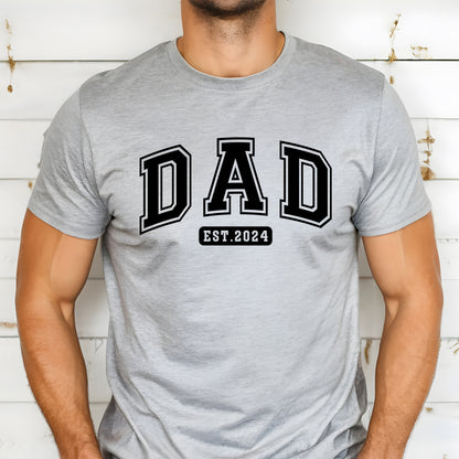 A stylish light colored t-shirt featuring the text "Dad Est. 2024" in a collegiate, sporty font, perfect for new fathers. Made from soft, breathable cotton, this shirt is an ideal gift for Father's Day or to celebrate a newborn's arrival. Available in multiple colors and sizes. Bella+Canvas 3001 t-shirt. ReadyShirtAim.com