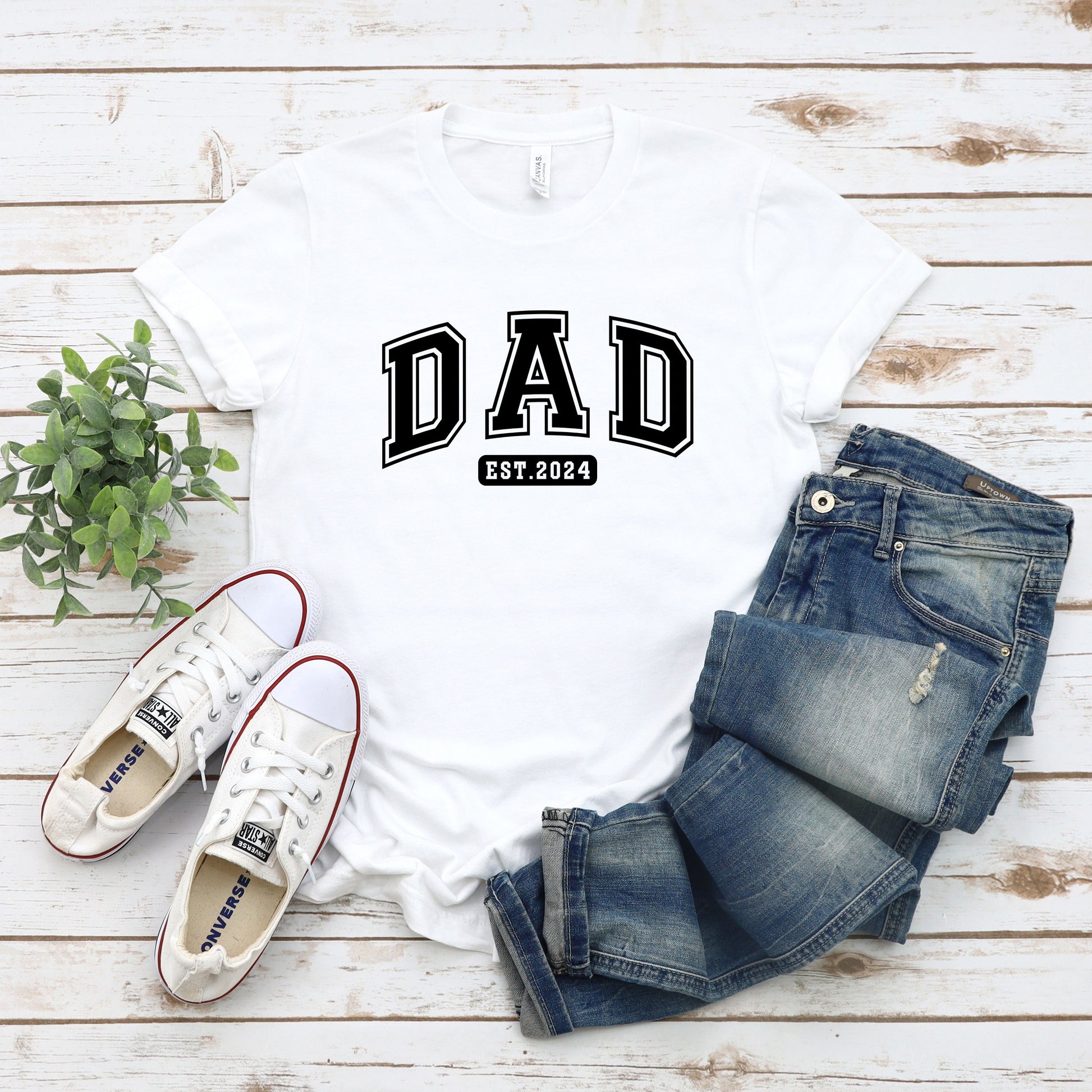 A stylish light colored t-shirt featuring the text "Dad Est. 2024" in a collegiate, sporty font, perfect for new fathers. Made from soft, breathable cotton, this shirt is an ideal gift for Father's Day or to celebrate a newborn's arrival. Available in multiple colors and sizes. Bella+Canvas 3001 t-shirt. ReadyShirtAim.com