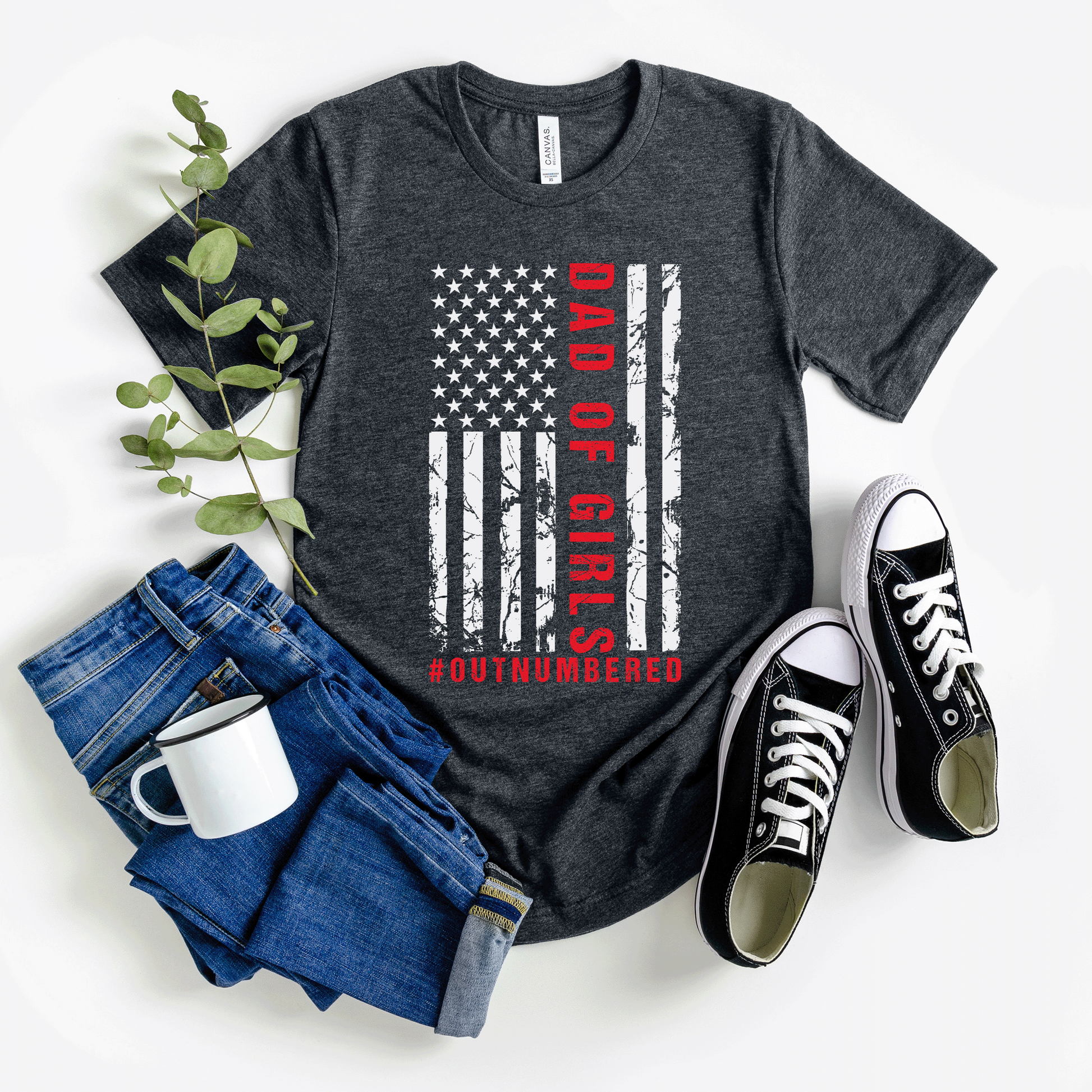 A dark-colored t-shirt featuring a grungy American flag design with the text "Dad of girls #outnumbered" in white and red. Perfect for Father's Day, Fourth of July, and patriotic dads who love humor. Comfortable and stylish for casual wear. Bella+Canvas 3001 t-shirt in dark grey heather. ReadyShirtAim.com