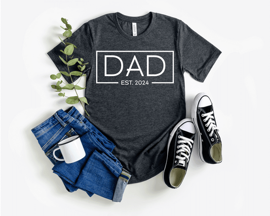 A dark t-shirt featuring the text "Dad Est. 2024" in a modern font, perfect for new fathers. Made from soft, breathable cotton, this shirt is an ideal gift for Father's Day or to celebrate a newborn's arrival. Available in multiple colors and sizes. Bella+Canvas 3001 t-shirt. ReadyShirtAim.com