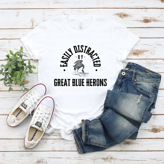 A vintage typeface t-shirt with a hand-drawn sketch of a great blue heron and the caption "Easily Distracted by Great Blue Herons." Perfect for heron lovers, bird watchers, and wildlife enthusiasts. Bella+Canvas 3001 t-shirt in white. ReadyShirtAim.com