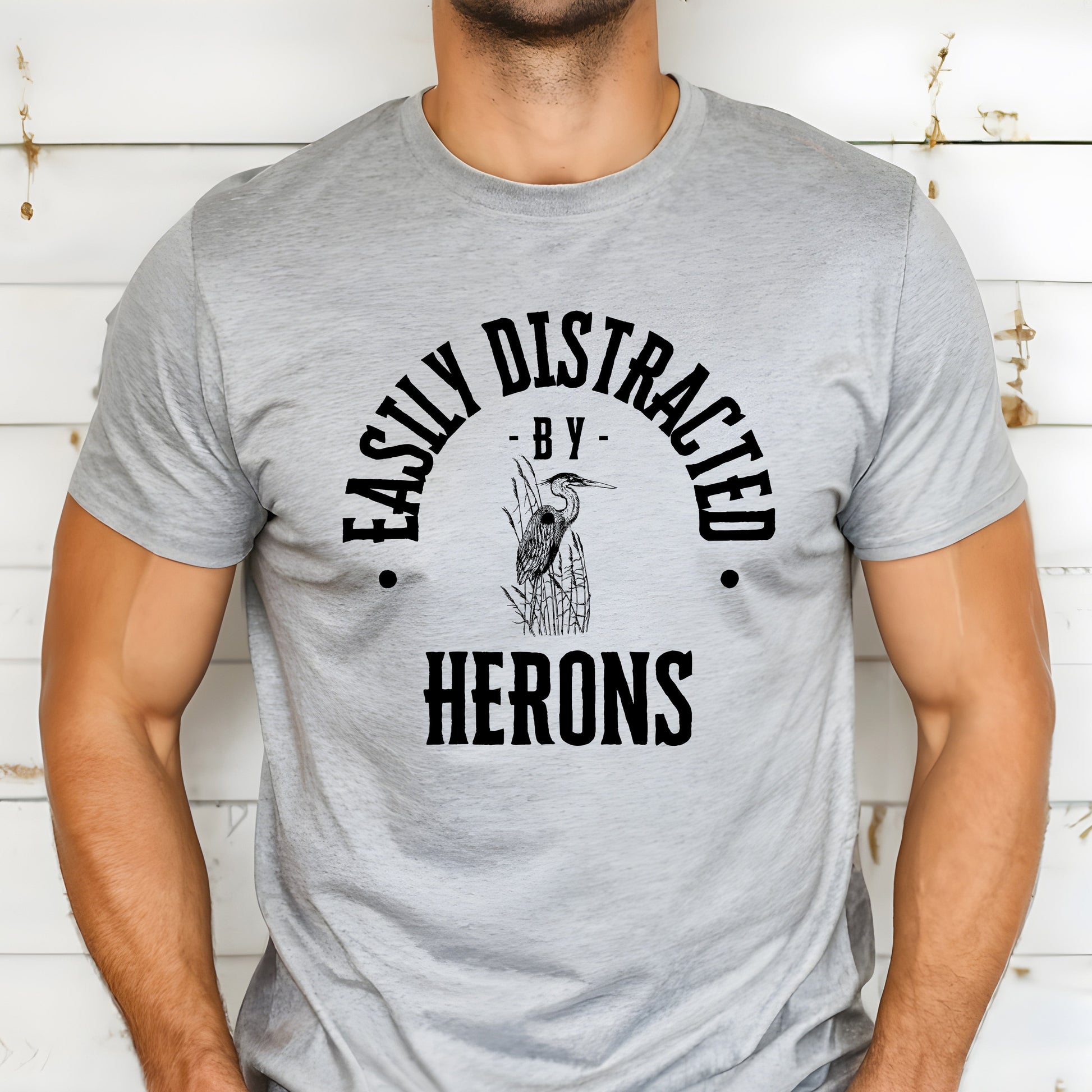 A vintage typeface t-shirt with a hand-drawn sketch of a heron and the caption "Easily Distracted by Herons." Perfect for heron lovers, bird watchers, and wildlife enthusiasts. Bella+Canvas 3001 t-shirt in athletic heather. ReadyShirtAim.com