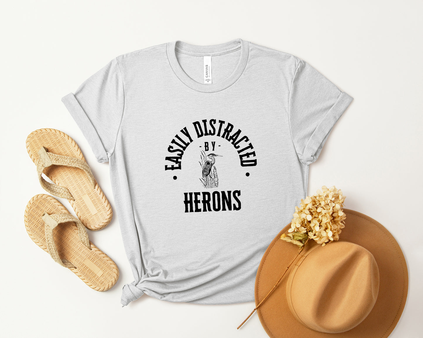 A vintage typeface t-shirt with a hand-drawn sketch of a heron and the caption "Easily Distracted by Herons." Perfect for heron lovers, bird watchers, and wildlife enthusiasts. Bella+Canvas 3001 t-shirt in ash. ReadyShirtAim.com