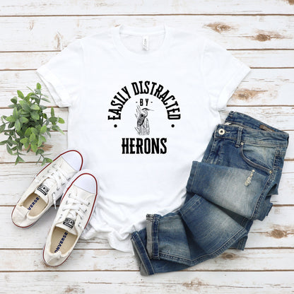 A vintage typeface t-shirt with a hand-drawn sketch of a heron and the caption "Easily Distracted by Herons." Perfect for heron lovers, bird watchers, and wildlife enthusiasts. Bella+Canvas 3001 t-shirt in white. ReadyShirtAim.com