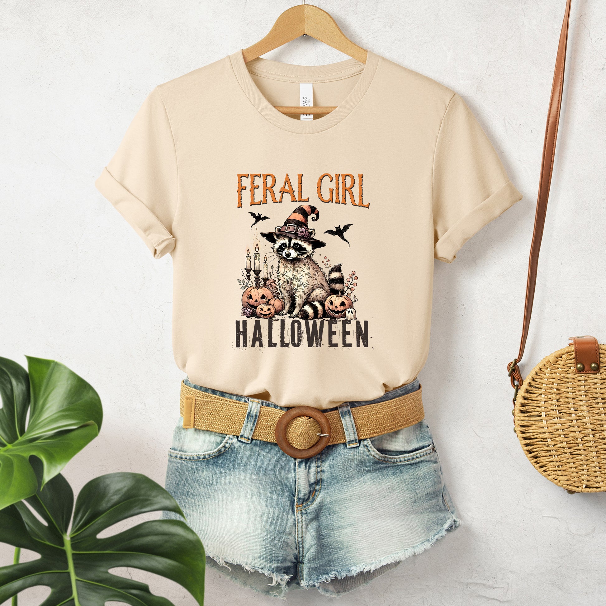 "Feral Girl Halloween" Funny Raccoon Witch Halloween Women's T-Shirt featuring dark brown text and a neutral-toned graphic of a raccoon dressed as a witch, smiling while sitting next to jack-o'-lanterns, surrounded by Halloween elements and bats flying in the air, perfect for adding a humorous touch to your Halloween festivities.