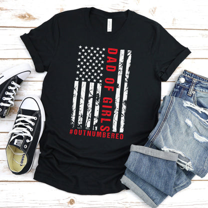 A dark-colored t-shirt featuring a grungy American flag design with the text "Dad of girls #outnumbered" in white and red. Perfect for Father's Day, Fourth of July, and patriotic dads who love humor. Comfortable and stylish for casual wear. Bella+Canvas 3001 t-shirt in black. ReadyShirtAim.com