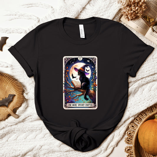 "One More Spooky Chapter" Black Cat Tarot Card Halloween Women's T-Shirt featuring a black cat in a witch's hat reading a book on a tree branch under a full moon, with bats flying around, in vibrant shades of orange, blues, and purples, perfect for adding a magical touch to your Halloween festivities. Available in multiple colors and sizes.  Bella+Canvas 3001 t-shirt in black. ReadyShirtAim.com