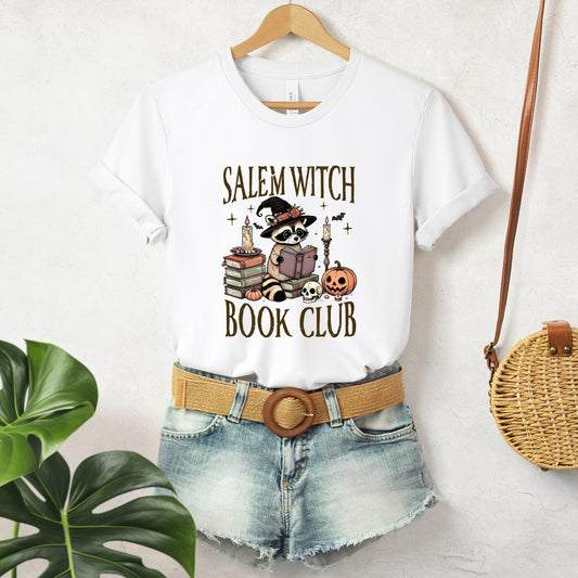 "Salem Witch Book Club" Cute Raccoon Halloween Women's T-Shirt featuring dark brown text and a neutral-toned graphic of a raccoon dressed in a witch's hat and reading glasses, reading a book, surrounded by jack-o'-lanterns, candles, skulls, and bats flying in the background, perfect for adding a whimsical touch to your Halloween festivities.