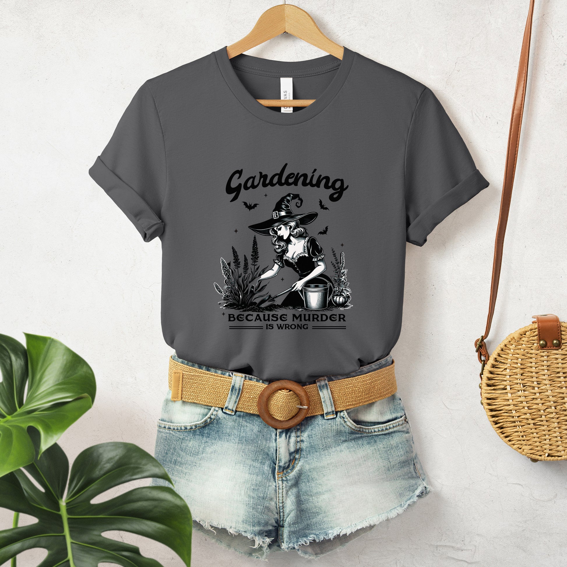 "Gardening, Because Murder is Wrong" Vintage 1950's Witch Halloween Women's T-Shirt featuring bold black text and a classic black-and-white image of a pin-up girl in a witch costume tending to flowers in a garden, smiling innocently, perfect for adding a retro touch with a playful twist to your Halloween festivities. Available in multiple colors and sizes.  Bella+Canvas 3001 t-shirt in asphalt. ReadyShirtAim.com