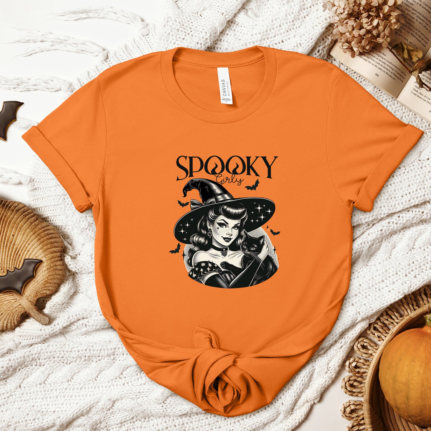 "Spooky Girly" Vintage 1950's Witch Halloween Women's T-Shirt featuring bold black text and a classic black-and-white image of a pin-up girl in a sexy witch costume, holding a black cat and smiling mischievously, perfect for adding a retro touch with playful charm to your Halloween festivities. Available in multiple colors and sizes.  Bella+Canvas 3001 t-shirt in orange. ReadyShirtAim.com