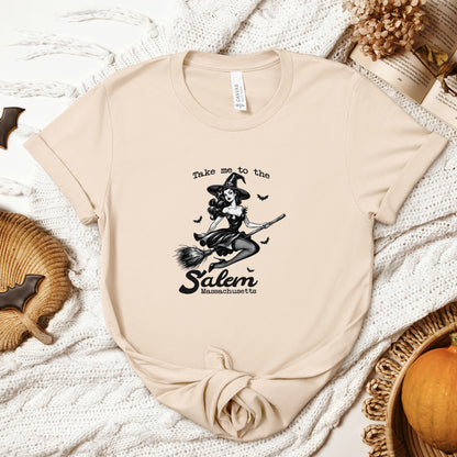 "Take Me to Salem, Massachusetts" Vintage 1950's Witch Halloween Women's T-Shirt featuring bold black text and a classic black-and-white image of a pin-up girl in a sexy witch costume flying gracefully on a broomstick with bats surrounding her, perfect for adding a retro touch with whimsical magic to your Halloween festivities. Available in multiple colors and sizes.  Bella+Canvas 3001 t-shirt in soft cream. ReadyShirtAim.com